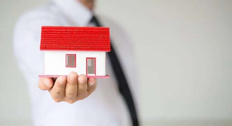 Un hombre con corbata negra sostiene una maqueta de casa blanca con techo rojo. ¿Listo para que puedas alquilar mi vivienda habitual?