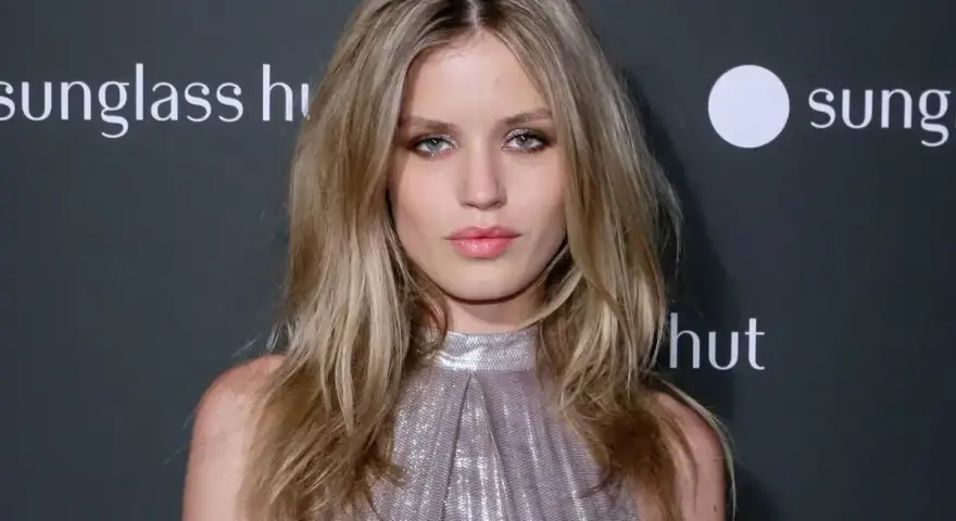 Modelka z chłodnymi odcieniami blondu, w błyszczącej sukience, pozuje na tle logo Sunglass Hut.