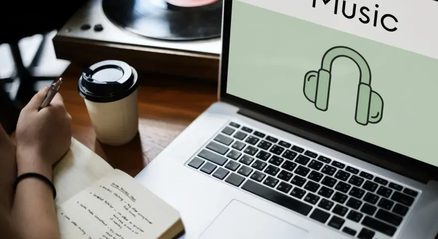 Osoba notuje pomysły na biznes, słuchając muzyki przez słuchawki. Laptop wyświetla ikonę słuchawek i napis "Music", sugerując rozpoznawanie muzyki.