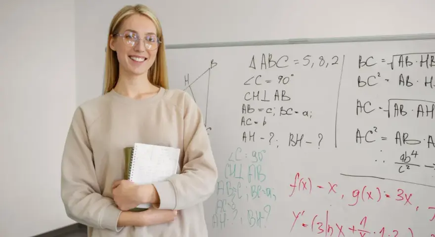 Uśmiechnięta nauczycielka z zeszytem przy tablicy z matematycznymi wzorami. Zastanawia się, ile punktów na maturze z matematyki można zdobyć za takie zadania.