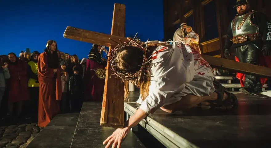 Scena drogi krzyżowej: Jezus w cierniowej koronie, z krwią na szacie, upada pod ciężarem krzyża. Tłum widzów obserwuje to poruszające przedstawienie.