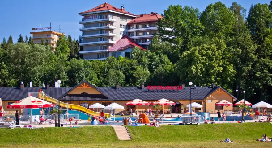 Słoneczny dzień w Muszynie-Zdroju. Ludzie relaksują się przy basenie zjeżdżalnią, a w tle widać hotel i restaurację.