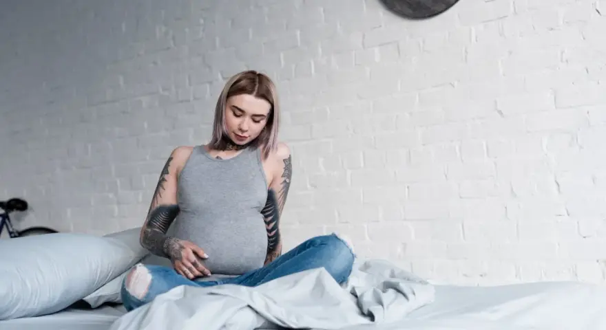 Donna incinta con tatuaggi sulle braccia e sul collo, seduta sul letto, con un pancione visibile.