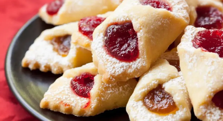 Ciastka kruche con ripieno di marmellata, spolverati di zucchero a velo, su un piatto scuro.