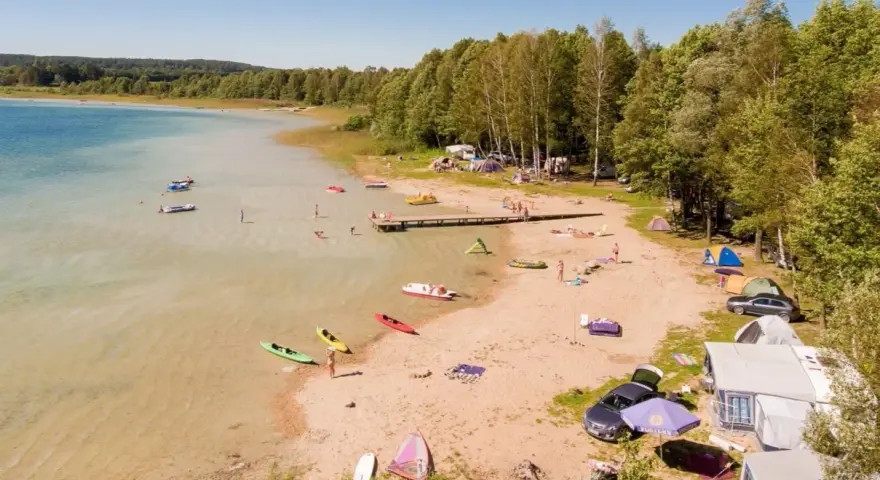Letnie popołudnie nad jeziorem Świętajno. Ludzie kąpią się, pływają kajakami i odpoczywają na plaży wśród namiotów i kamperów.