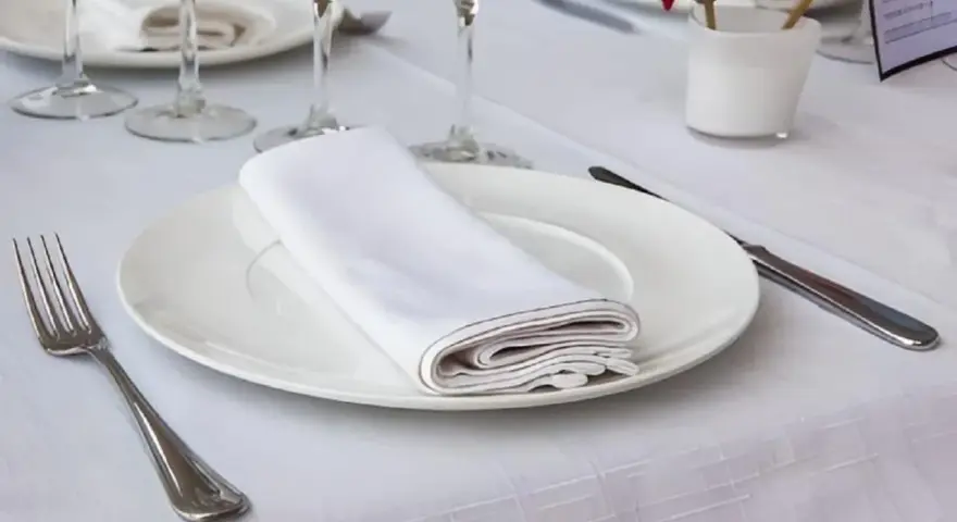 Une table dressée avec une assiette blanche, une serviette de table pliée, une fourchette et un couteau. La dimension serviette de table est parfaite pour cette présentation élégante.