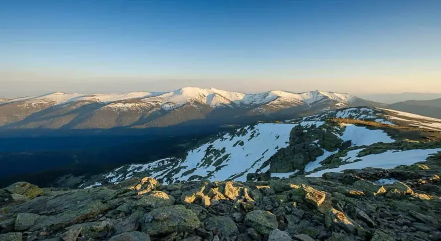 Paisaje montañoso nevado al amanecer, con rocas en primer plano y el pico Madrid en la distancia.