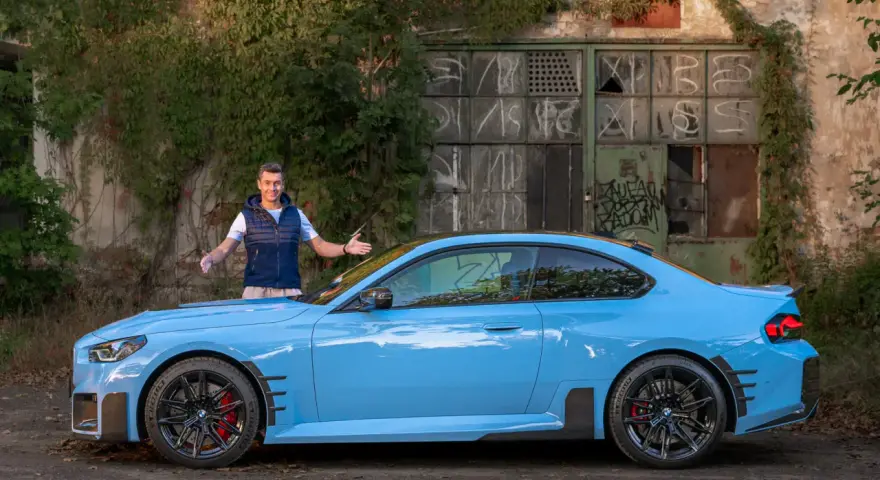 Mężczyzna prezentuje błękitne BMW M2 G87 przed zniszczonym budynkiem.