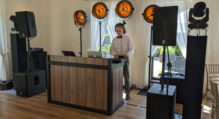 Młody DJ przy konsoli, otoczony sprzętem nagłośnieniowym i oświetleniem. Unikaj błędów w wyborze oprawy muzycznej – DJ czy zespół?