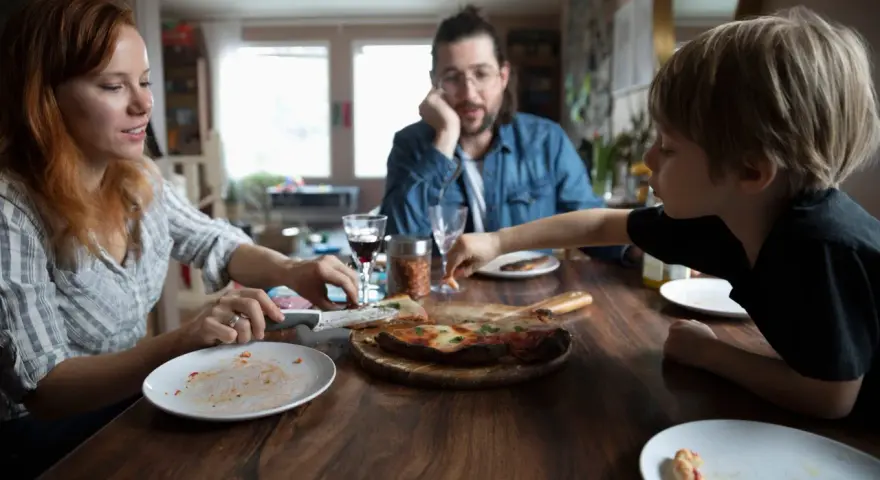 Rodzina przy stole, pizza, w tle mężczyzna zamyślony. Czasem nawet przy wspólnym posiłku pojawiają się kłótnie o dzieci z poprzedniego związku.