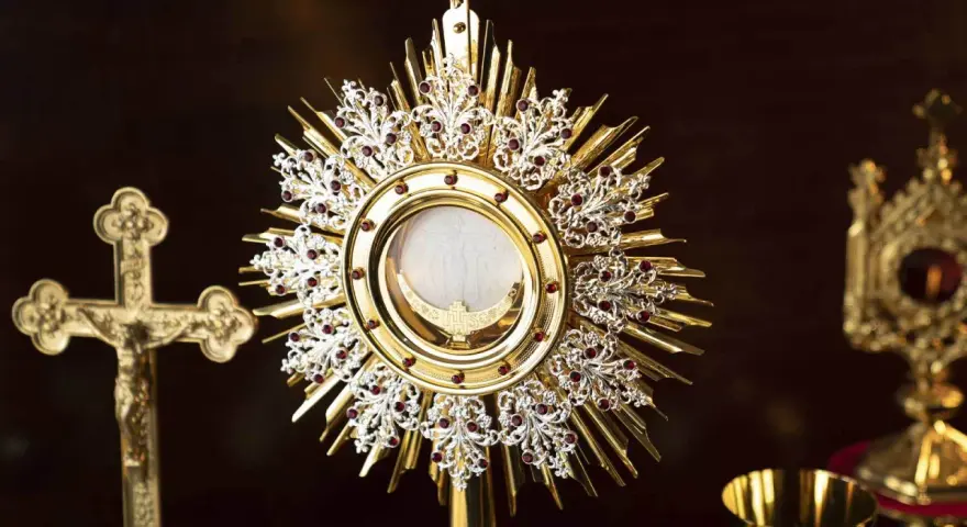 A golden monstrance holds the blessed sacrament, surrounded by ornate details and red jewels. A crucifix stands nearby, a symbol for a blessed sacrament prayer request.