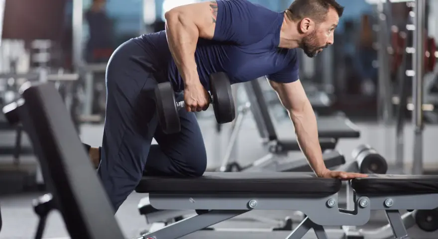 Man utför enarms hantelrodd på ett gym. Han stöttar sig mot en bänk med ena handen och drar en hantel mot bröstet med den andra.