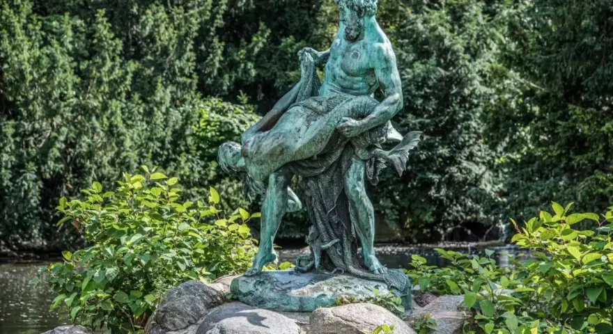 Bronzestatue eines Mannes, der eine Frau trägt, vor einem grünen Hintergrund mit einem kleinen Wasserfall in Berlin.