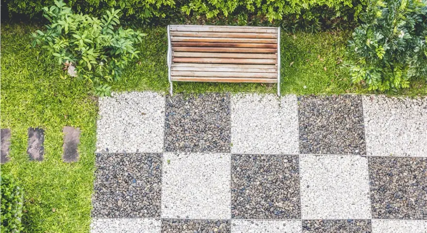 Ideas de suelo para terraza: un banco de madera sobre césped y un patrón de tablero de ajedrez de guijarros blancos y grises.