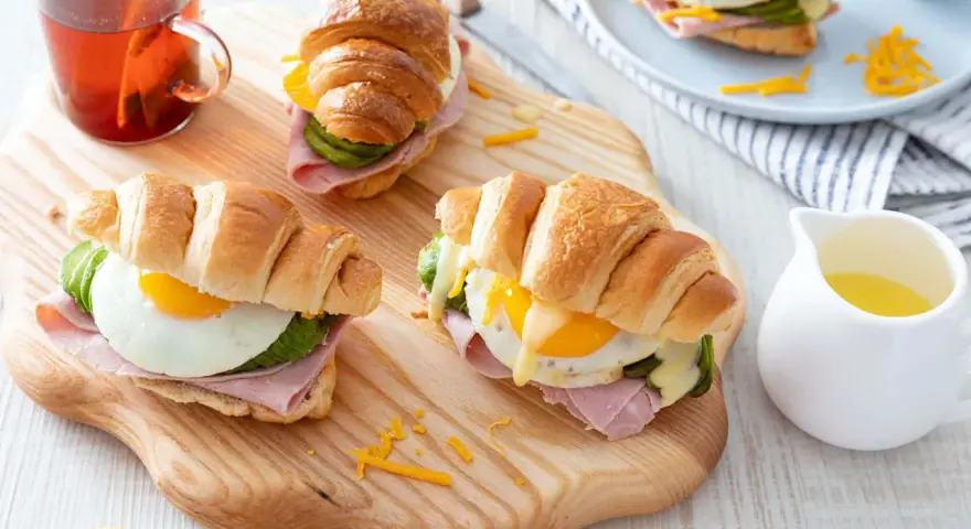 Trois croissants au jambon, œuf au plat, avocat et fromage râpé, servis sur une planche en bois avec une tasse de thé.