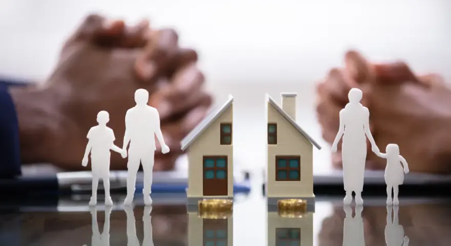 Figurines de famille séparées par une maison coupée en deux, symbolisant une convention d'indivision divorce. Mains floues en arrière-plan.