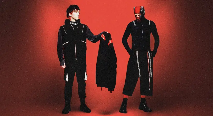 Dwóch artystów w czarnych strojach, przypominających styl z okładek płyt twenty one pilots, pozuje na tle czerwonym.