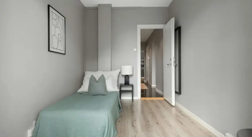 Habitación minimalista con cama individual, mesita de noche y lámpara. La puerta abierta revela un pasillo, sugiriendo que es legal alquilar habitaciones.
