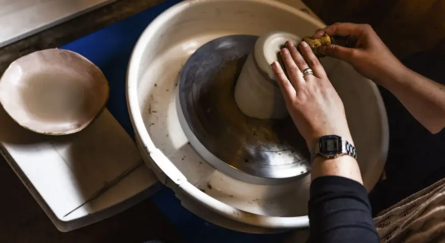 Händer som formar en mugg på en drejskiva. En svamp används för att jämna till leran medan man drejar lera.