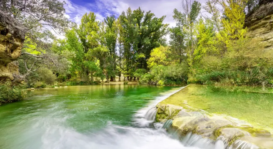 La poza de doña urraca, un remanso de agua verde esmeralda con una pequeña cascada, rodeada de frondosa vegetación y árboles altos bajo un cielo azul.