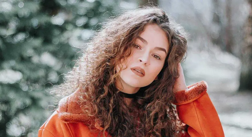 Jeune femme aux cheveux bouclés, portant une veste orange. La retouche photo netteté en ligne sublime son regard intense.