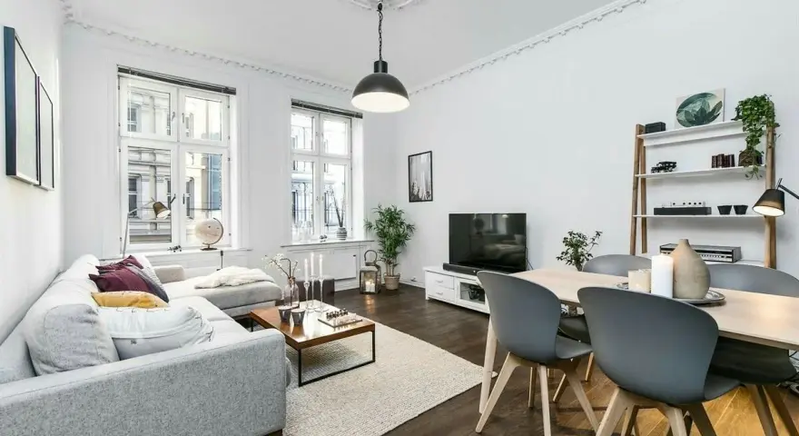 Elegante salón con sofá gris, mesa de centro y comedor. El **suelo oscuro** de madera contrasta con las paredes blancas y la luz natural.