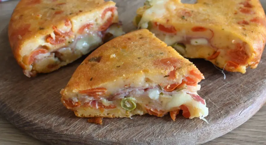 Fette di torta salata senza uova, ricca di pomodoro, formaggio filante e prosciutto, su un tagliere di legno.