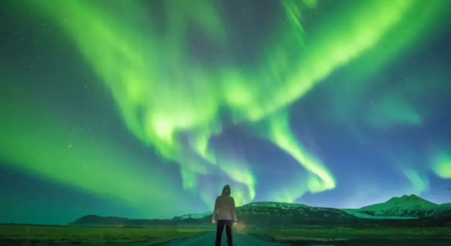 Człowiek stoi na drodze pod rozgwieżdżonym niebem, gdzie tańczy zorza polarna. Islandia, październik, pogoda idealna na takie widowisko.