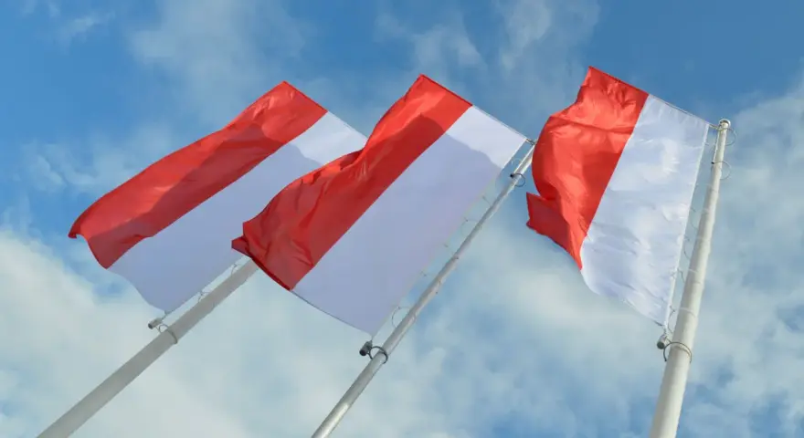 Trzy powiewające na wietrze flagi, każda z czerwono-białą szarfą, na tle błękitnego nieba z chmurami.