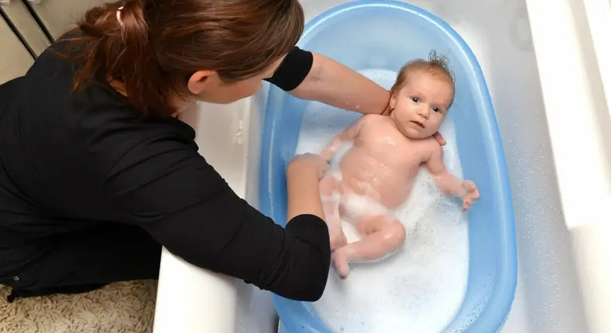 Mama delikatnie myje noworodka w niebieskiej wanience. Maluch pluska się w pianie, a mama uśmiecha się do niego.