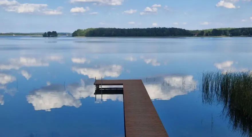 Drewniane molo na spokojnym jeziorze Wysockim, odbijającym błękitne niebo z białymi chmurami.