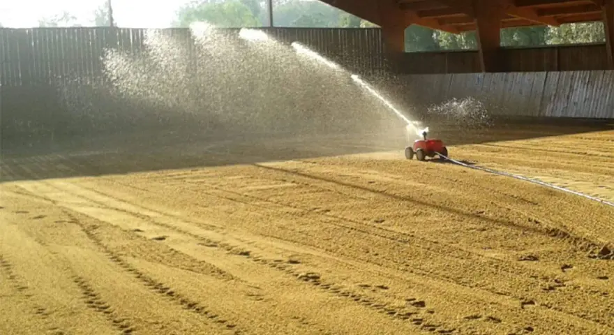 Arrosage d'une carrière équestre par un système mobile, créant un voile d'eau sur le sable.
