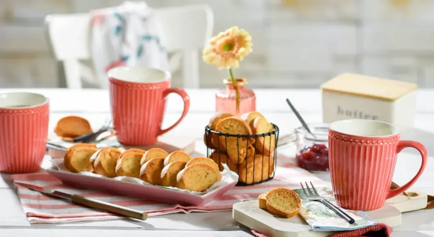 Leckeres Zwieback-Rezept: goldbraune Scheiben auf rosa Platte, dazu Marmelade, Butterdose und rote Tassen.