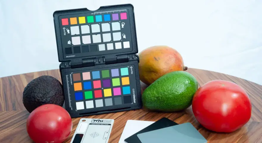 Na drewnianym blacie leżą owoce: awokado, mango, pomidory. Obok znajduje się otwarty colorchecker z kolorowymi polami i szara karta do kalibracji balansu bieli.