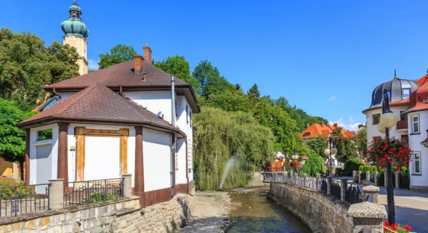 Urokliwy strumyk w Polanicy Zdrój, otoczony kamiennymi murkami i zielenią. W tle widać wieżę kościelną i kolorowe kamieniczki.