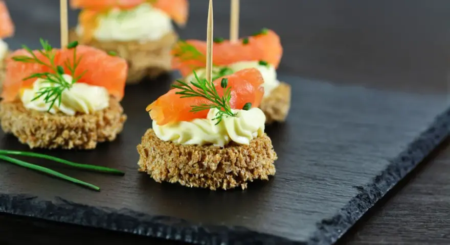 Eleganckie przystawki na komunię: krążki ciemnego pieczywa z kremowym serkiem, łososiem i koperkiem.