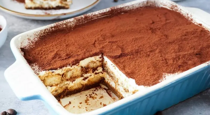 Ein köstliches Tiramisu rezept ohne alkohol, bestäubt mit Kakao, in einer blauen Auflaufform. Ein Stück ist bereits entnommen.