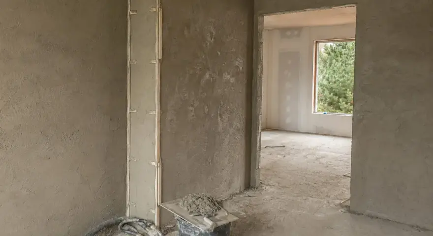 Wnętrze w trakcie prac wykończeniowych, ściany pokryte tynkami gipsowymi lub cementowo wapiennymi. Widoczne wiadro z zaprawą i narzędzia.