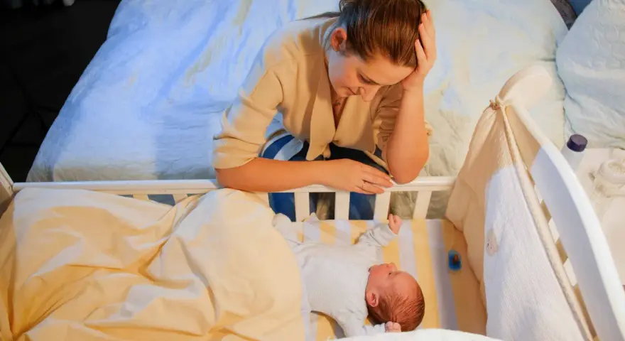 Zmęczona mama pochyla się nad łóżeczkiem, zastanawiając się, kiedy nastąpi regres snu. Niemowlę śpi spokojnie.