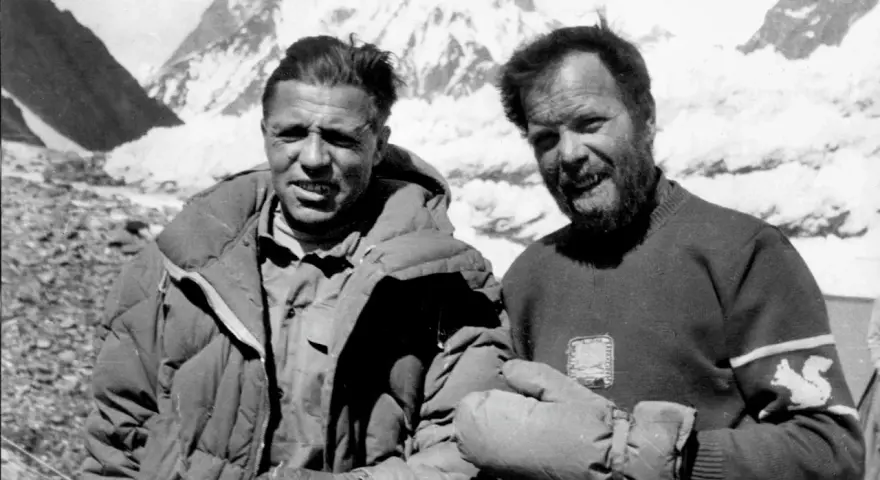 Zwei Bergsteiger, die die K2 Erstbesteigung schafften, lächeln in die Kamera. Im Hintergrund sind schneebedeckte Berge zu sehen.