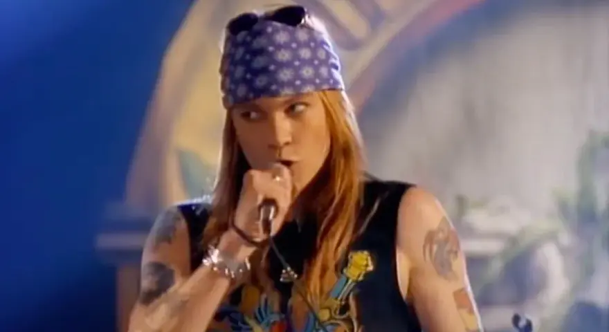 Axl Rose w fioletowej bandanie i czarnej koszulce śpiewa do mikrofonu, jakby dedykował "sweet child o' mine" publiczności.