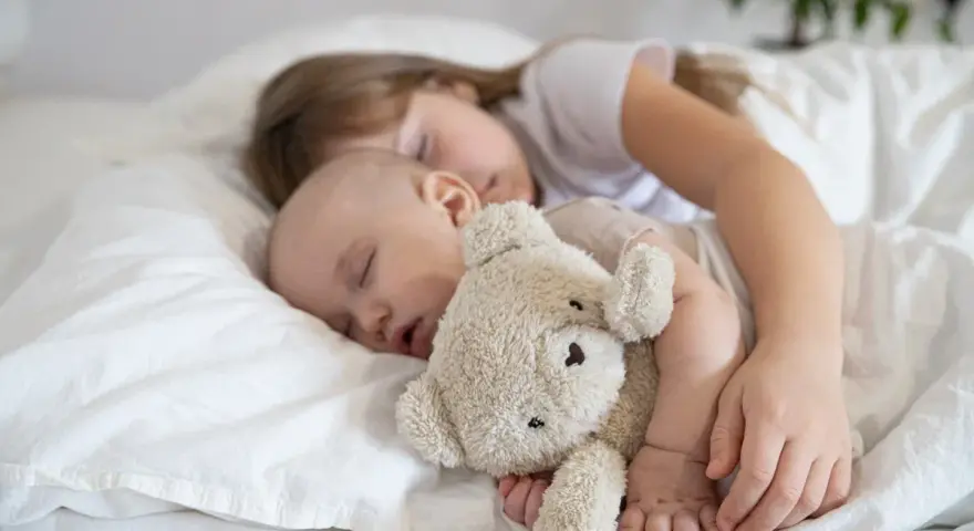 Dzieci śpią wtulone w misia. Starsza siostra czuwa nad maluszkiem, pokazując, ile śpi dwulatek i jak ważny jest sen dla rozwoju.