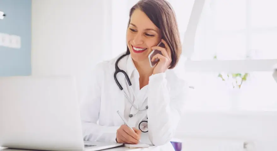 Lekarka w białym kitlu rozmawia przez telefon i pisze notatki przy laptopie.