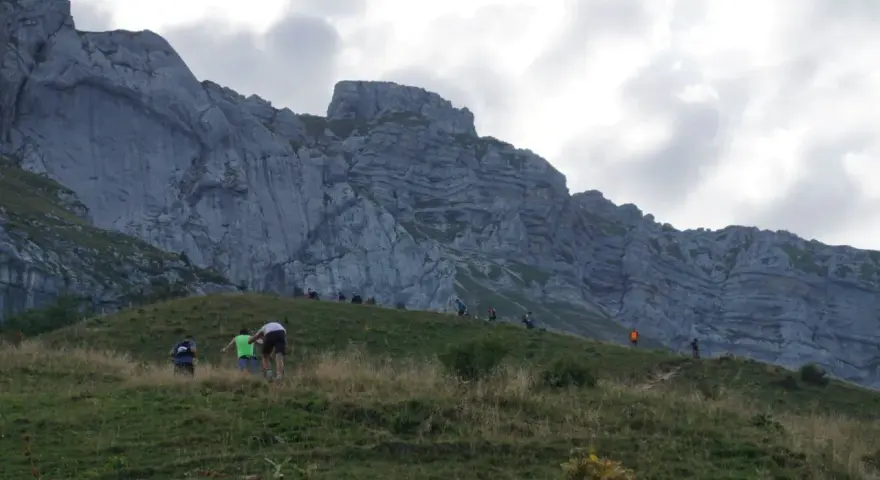 Randonneurs sur une pente herbeuse face à une falaise imposante. Le chemin sinueux et la hauteur suggèrent que la tournette est dangereuse.