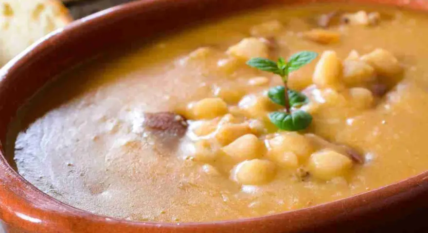 Una ciotola di terracotta piena di zuppa di fagioli cremosa, con pezzetti di carne e una fogliolina di menta. Perfetta per una cena confortante.