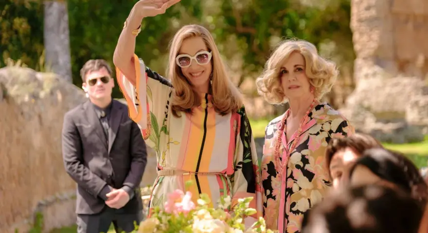 Due donne eleganti, una saluta con la mano, in un giardino. Un uomo in giacca scura fa da guardia. Un semplice favore per un evento speciale.