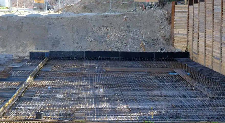 Armatura per fondazione a platea sezione. La rete metallica è pronta per il getto di calcestruzzo, con terra e muri di contenimento sullo sfondo.