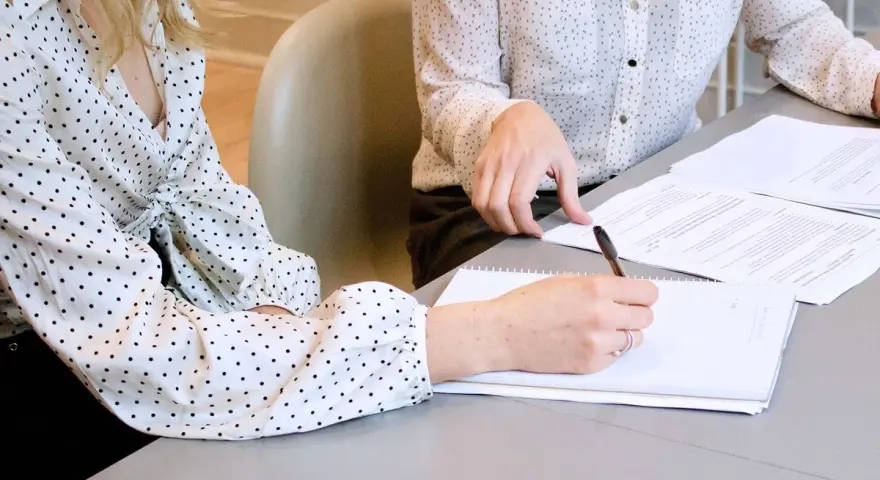 Two women in polka-dot shirts review documents. One takes notes, implying they are discussing who can see committee meeting minutes.