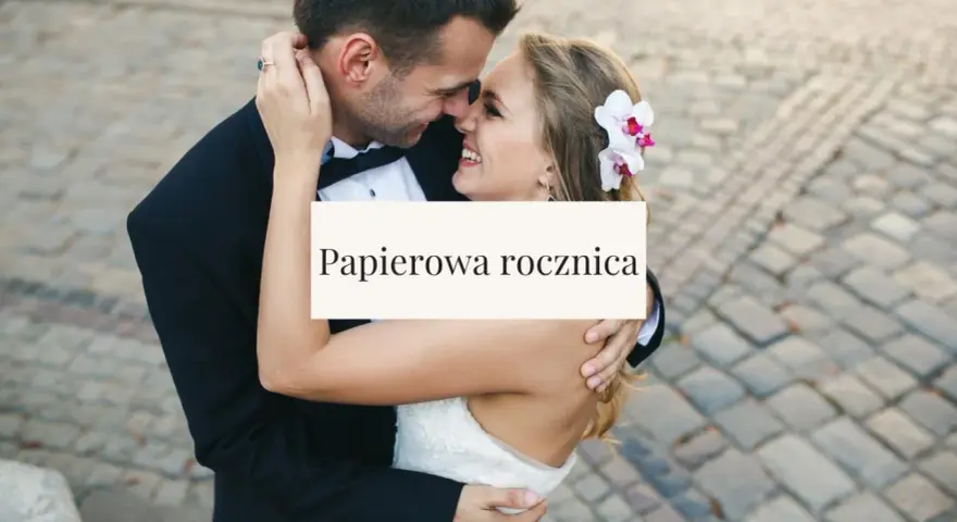 Para świętuje papierową rocznicę ślubu. Uśmiechnięci, w objęciach, z kwiatem we włosach panny młodej.
