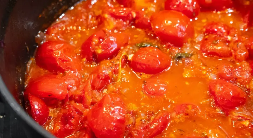 Ein köstliches, blubberndes Tomaten Sugo in einer schwarzen Pfanne, voller saftiger Kirschtomaten und aromatischer Kräuter.